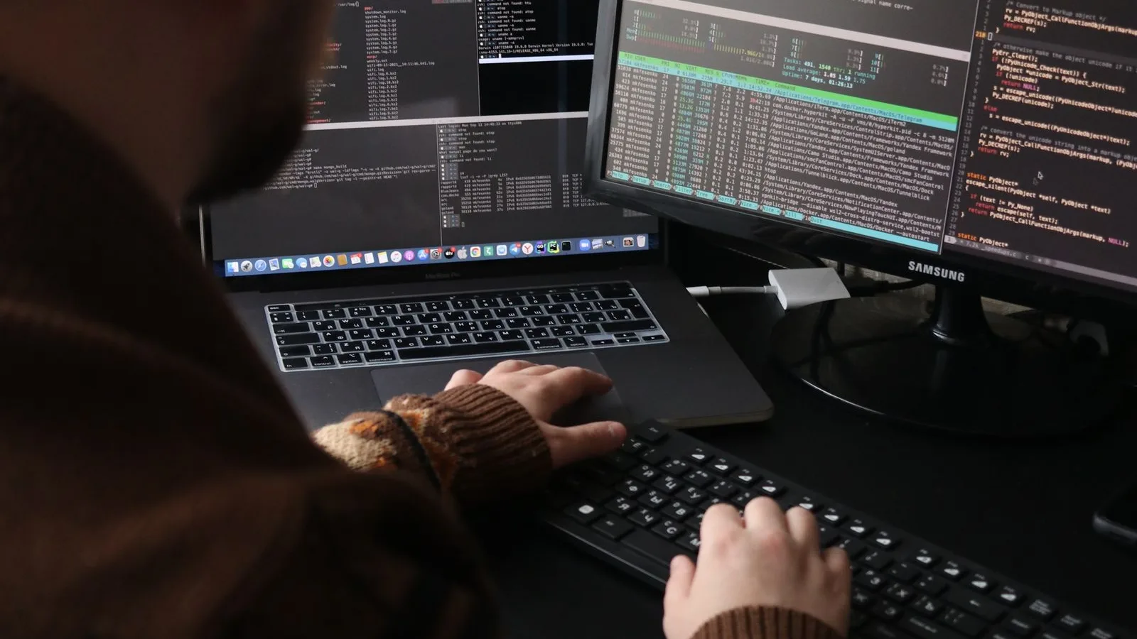 A programmer working on code with dual monitors and a laptop in a dimly lit room